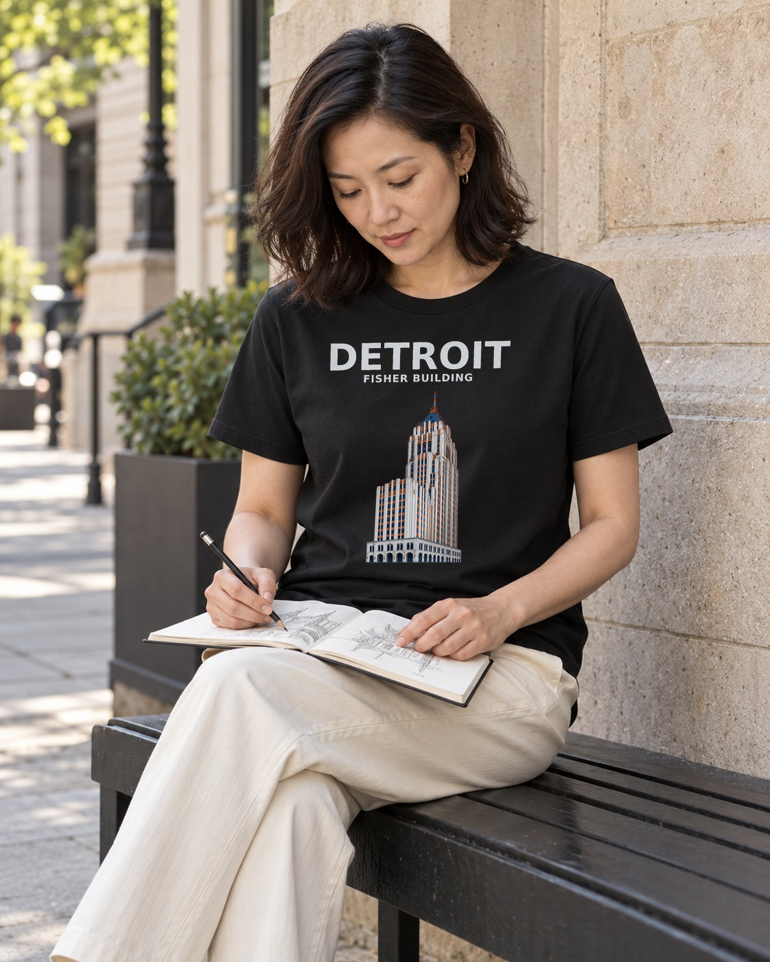 Illustrative lifestyle mockup of the black Detroit Fisher Building T-shirt, with a person sketching in a generated city scene.